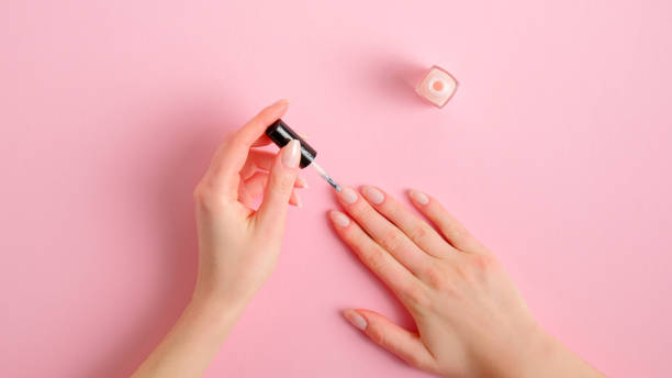 Woman applying polish on nails at home. Female hands with elegant manicure and nail polish bottle on pink background, top view. Beauty treatment and hand care concept stock photo
