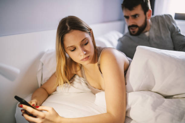 istock Young attractive blonde lying in the bed and using smart phone for replying messages to her lover. In background is her jealous boyfriend arguing with her. 1208105762