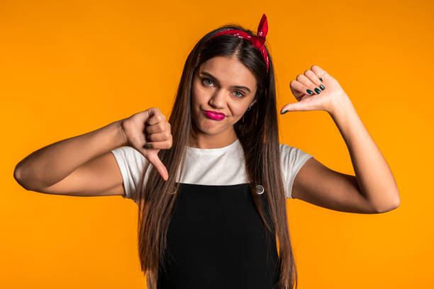 istock Young pretty woman standing on yellow studio background expressing discontent and showing thumb down gesture at camera. Portrait of girl with sign of dislike 1208112994
