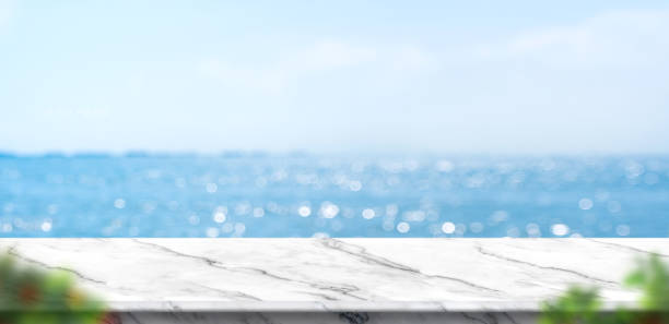 Empty white marble table with blur blue sky and sea bokeh background with green leaf foreground,Mock up template for display or montage of product or content use as banner in social media ads mesa de mármol blanco vacía con el cielo azul borroso y fondo bokeh mar con el primer plano de la hoja verde, plantilla de la punción para la visualización o montaje del producto o el contenido del uso como banner en los anuncios de las redes sociales