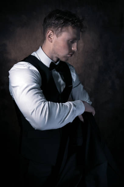 istock Good looking man with beard, curly hair wearing waistcoat and bowtie pulling on his jacket, looking away from camera 1208119602