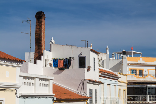 Mostly white facades of the houses with orange or flat roofs. Chimney in between of them. Light clouds on a blue sky. Alvor, Algarve, Portugal.