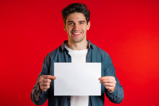 istock Handsome man holding horizontal white a4 paper poster. Copy space. Smiling hipster guy in shirt on red background. 1208129635