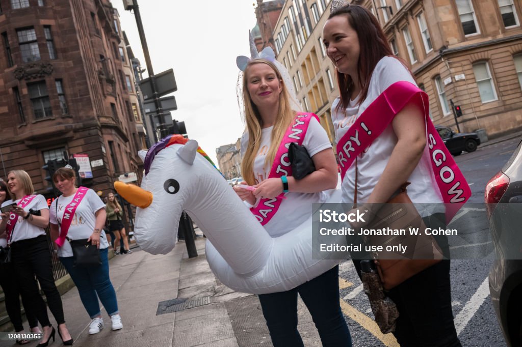 A bride, wearing a unicorn head piece with a veil and unicorn floatation device, walks the streets of Glasgow with her friends at her hen-party, prior to her wedding. Glasgow, Scotland/UK, June 29, 2019: A bride, wearing a unicorn head piece with a veil and unicorn floatation device, walks the streets of Glasgow with her friends at her hen-party, prior to her wedding. Stag Night Stock Photo