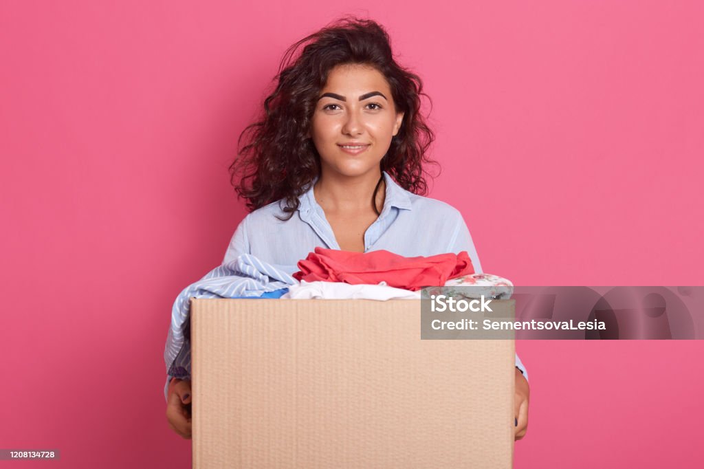 Image of dark haired adorable kind young woman standing isolated over pink background in studio, holding paper box with donated clothes, wearing blue shirt, doing good things. Charity concept. Adult Stock Photo