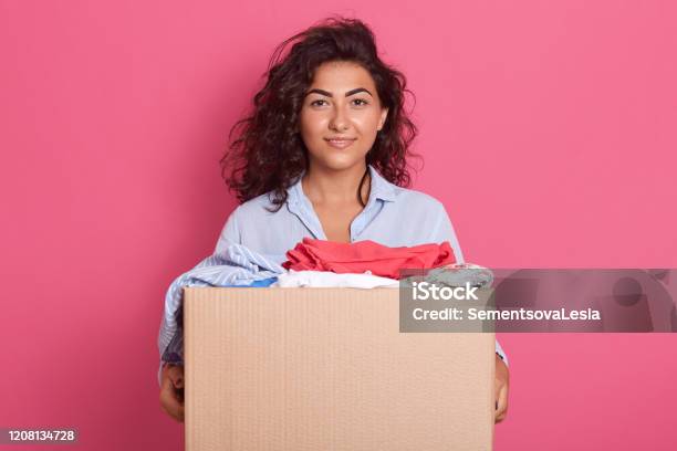 Image Of Dark Haired Adorable Kind Young Woman Standing Isolated Over Pink Background In Studio Holding Paper Box With Donated Clothes Wearing Blue Shirt Doing Good Things Charity Concept Stock Photo - Download Image Now