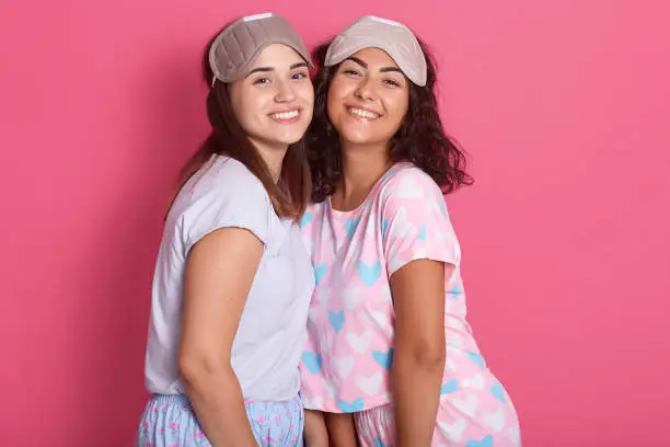 Photo of Horizontal shot of beautiful girls posing in pajamas and sleeping mask, girls friends standing issolated over pink background, having pajama party, likes spending time together. Friendship concept.