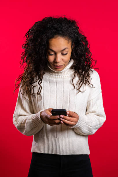 istock Girl with afro hair using smartphone, surfing internet or playing game on red studio background. Modern technology - apps, social networks 1208145134