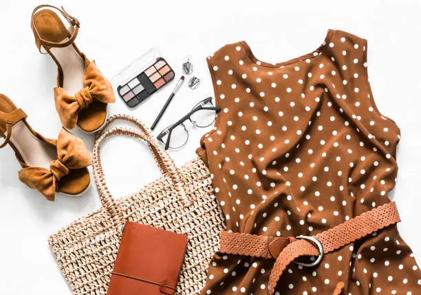 Photo of Polka dot summer brown dress, suede wedge sandals, eco straw tote bag, cosmetics on a light background, top view. Women's clothing set