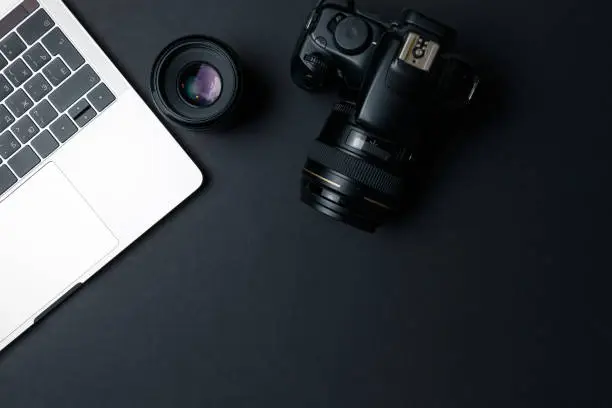 Photo of Photographer's workplace on a dark background. Modern laptop, digital camera, lens, battery, smartphone. Minimalism. Top view. Copy space. Equipment for the photographer. The concept of freelancing
