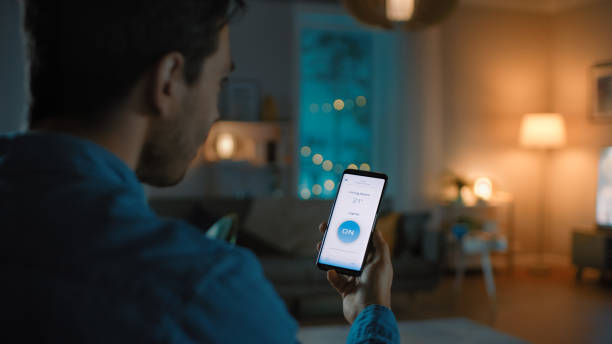 istock Young Handsome Man Gives a Voice Command to a Smart Home Application on His Smartphone To Switch on the Lights and Set a Comfortable Temperature. It's a Cozy Evening in Living Room. 1208159379