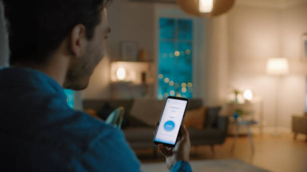 istock Young Handsome Man Gives a Voice Command to a Smart Home Application on His Smartphone To Switch on the Lights and Set a Comfortable Temperature. It's a Cozy Evening in Living Room. 1208159380
