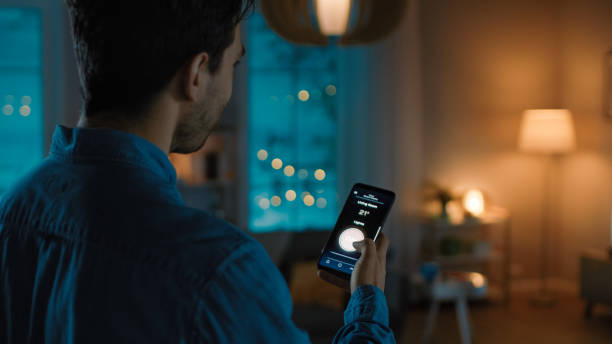 istock Young Handsome Man is Holding Smartphone with Active Smart Home Application. Person is Tapping the Screen to Turn On/Off the Lights in the Room. It's Cozy Evening in the Apartment. 1208159911