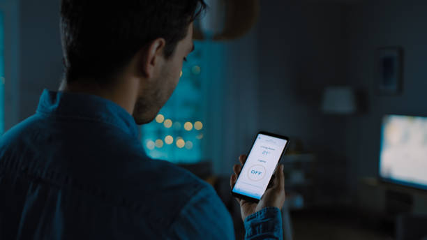 istock Young Handsome Man Gives a Voice Command to a Smart Home Application on His Smartphone To Switch off the Lights and Set a Comfortable Temperature. It's a Cozy Evening in Living Room. 1208159916