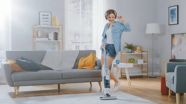shot of a young beautiful woman in jeans shirt and shorts dancing and vacuum cleaning a carpet in a cozy room at home. she uses a modern cordless vacuum. she's happy. - spring cleaning women cleaning dancing imagens e fotografias de stock