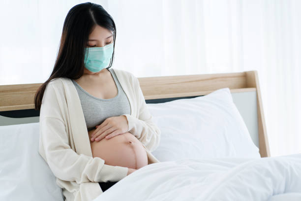 Pregnant Asian woman wearing a PM2.5 dust mask and Covic-19 (Coronavirus) is sitting on clean bed. Prevent infection to the fetus. Discomfort with flu. Should get enough rest and see a doctor. stock photo