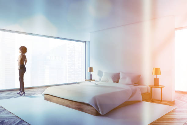 Young African American woman standing in luxury panoramic bedroom with white walls, wooden floor and comfortable king size bed. Toned image