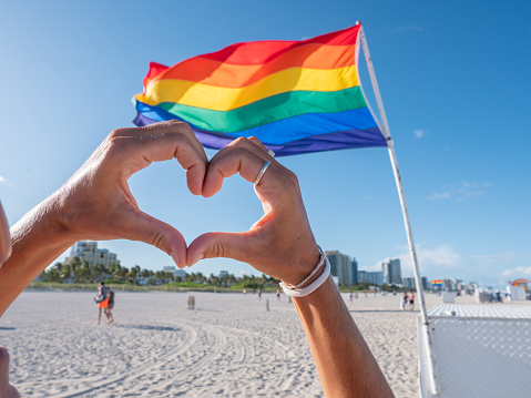 Cute girl on beach making heart shape with hands near rainbow gay flag on beach in Miami