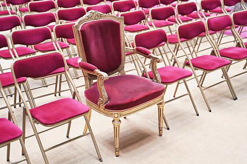 Trono único o sillón ceremonial con asiento de terciopelo y detalles dorados entre muchas sillas simples idénticas similares. Concepto de singularidad o exclusividad. photo