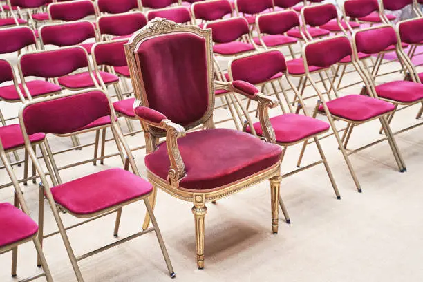 Photo of Unique throne or ceremonial armchair with velvet seat and golden details among many simple identical similar chairs. Uniqueness or exclusivity concept.