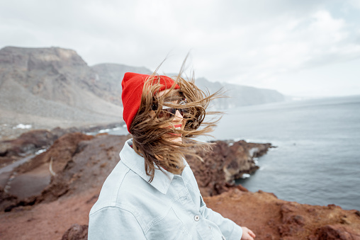 Lifestyle portrait of a stylish woman enjoying trip on a rocky ocean coast during a strong wind. Traveling on Tenerife island, Spain