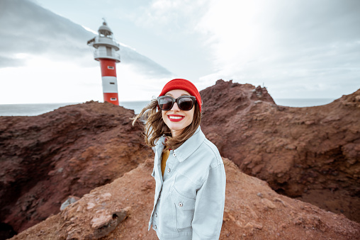 Portrait of a carefree stylish woman dressed in jeans and red hat enjoying trip on a rocky ocean shore near the lighthouse, traveling on north-west of Tenerife island, Spain