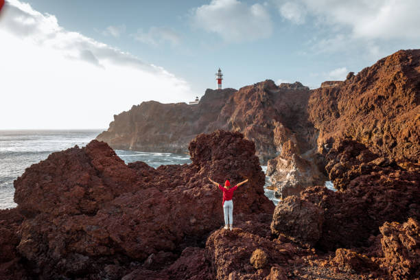 istock Woman on the rocky ocean coast with lighthouse on a sunset 1208193228