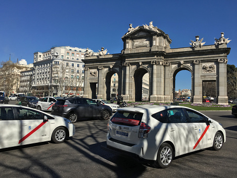Madrid, Spain - February 23, 2020: Official white taxis driving through the Calle de Alcala next to the Puerta de Alcala in the city downtown. This is a neo-classical monument commisisioned to Francesco Sabatini by King Charles III as a gate to the city walls that existed during that time