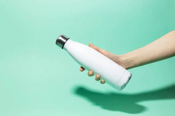 Close-up of female hand holding white reusable steel stainless thermo water bottle isolated on background of aqua menthe color. Plastic free.