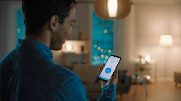 istock Young Handsome Man Gives a Voice Command to a Smart Home Application on His Smartphone To Switch on the Lights and Set a Comfortable Temperature. It's a Cozy Evening in Living Room. 1208201717