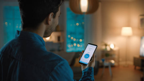 istock Young Handsome Man Gives a Voice Command to a Smart Home Application on His Smartphone To Switch on the Lights and Set a Comfortable Temperature. It's a Cozy Evening in Living Room. 1208202807