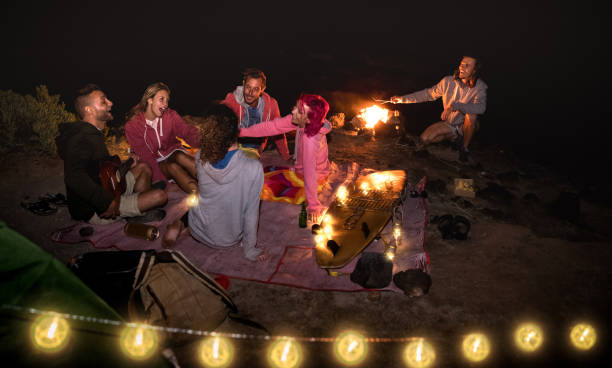 young hippie friends having fun together at night beach party with string light - friendship travel concept with young people traveler drinking beer at summer surf campfire - high iso image - bonfire beach fire barbecue imagens e fotografias de stock