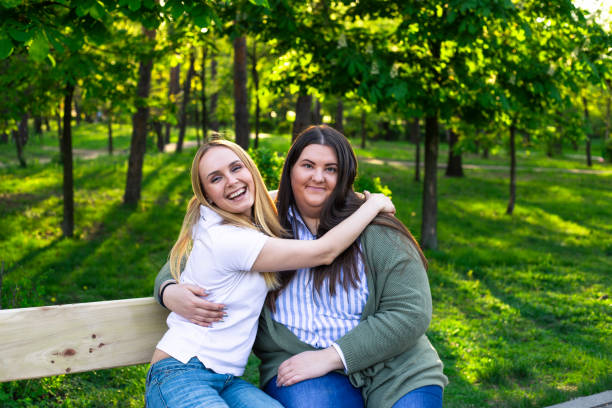 istock Two beautiful smiling young models sitting on the park bench and hugging sunny spring day. 1208214409
