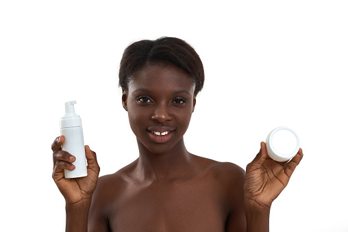 Portrait of beautiful young african woman demonstrating cosmetic products and looking at camera while standing in studio against white background. Skin care. Beauty concept. Cosmetology concept