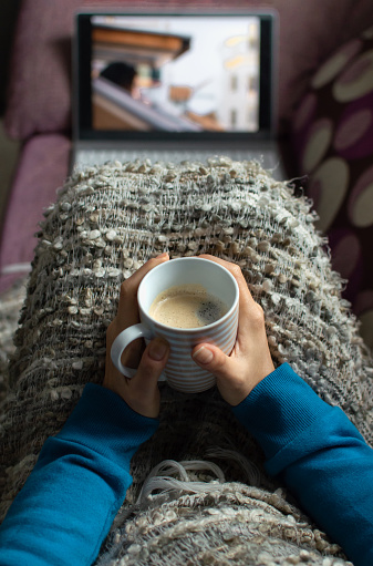 Woman sitting on a sofa with a coffee cup and covered with a blanket watching a serie or video on a laptop.