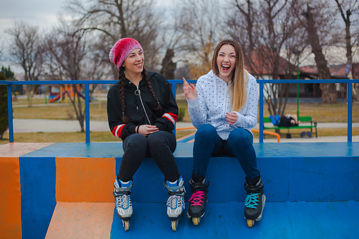 Two attractive young women on roller skates having fun while sitting in skate park.