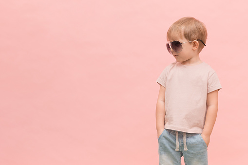 A blond European-looking child boy stands in sunglasses and looks left on a pink background. Concept with place for text, for articles about models, castings and fashion shows.