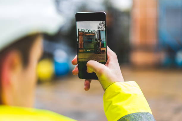 建設現場でスマートフォンを持っている建築家 - 若い建設労働者は、現場で携帯電話を使用しています - 建築計画と携帯電話を持つ建設労働者 - モバイルに焦点を当てています。暖かいフィ� - 建設 写真 ストックフォトと画像
