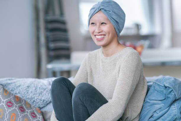 A courageous young Filipino woman is fighting cancer. She is wearing a blue headscarf to hide hair loss from chemotherapy treatment. The woman is sitting on a couch and has her arms wrapped around her legs. She is laughing and is filled with gratitude and hope for recovery.
