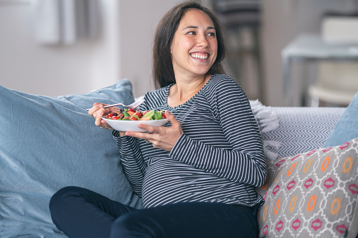 A pregnant woman of Eurasian descent is in her second trimester. The excited mother to be is enjoying a healthy meal at home. The woman is smiling while holding a salad. She is sitting on the couch in her living room.