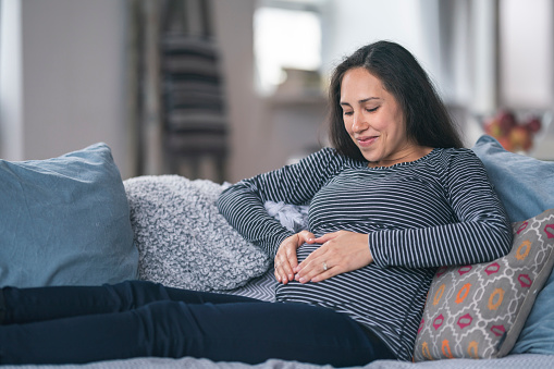 A young woman of Eurasian descent is pregnant. She is in her second trimester. The woman is relaxing on the couch in her home. She is smiling and looking down at her abdomen. She has formed the shape of a heart with her hands on her abdomen.