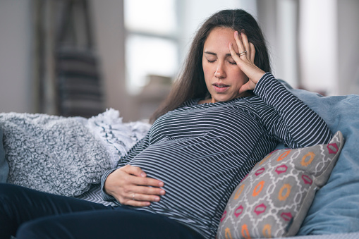 A pregnant Eurasian woman is in her second trimester. The woman is sitting on a couch and is holding her belly in discomfort. She is breathing heavily and is resting her head in her hand. Prenatal, morning sickness, back pain, heartburn, constipation, and fatigue concepts.