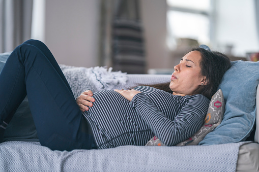 A young woman of Asian descent is pregnant. She is in her second trimester. The woman is lying on the couch in discomfort. She is breathing heavily and is resting her hand on her stomach. Prenatal, morning sickness, back pain, heartburn, and constipation concepts.