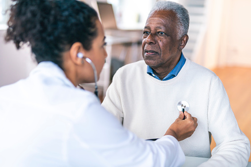 A black senior adult man is at a routine medical appointment. He is seated facing his female doctor. The doctor is using a stethoscope to check the man's heart and lungs. The image is shot over the shoulder of the medical professional and is focused on the patient.