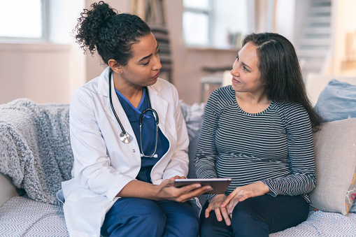 A female physician is meeting with a patient. The patient is a mixed-race pregnant woman in her second trimester. The two women are having a pleasant conversation. The doctor is holding a wireless tablet computer. The pregnant woman is smiling and listening to her doctor's advice.