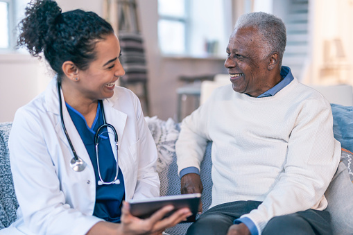 A female physician is meeting with a patient in the patient's home. The patient is a black senior adult man. The doctor and patient are seated next to each other on a couch. The doctor is holding a wireless tablet computer. The two individuals are smiling and looking at each other.