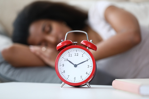 Close-up of red mechanical alarm clock standing on bedside table showing ten minutes to eleven. Young latino woman enjoying sweet dream on background. Wake up concept