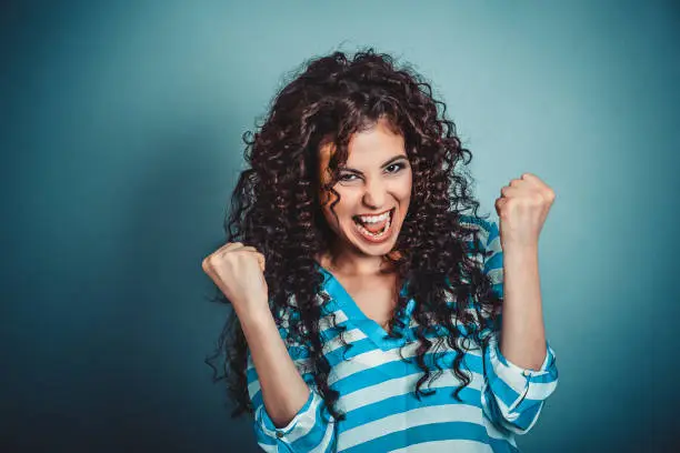 Photo of Lucky girl. Closeup portrait happy young brunette curly woman happy exults pumping fists ecstatic isolated blue background. Celebrate success concept. Human facial expression emotions body language
