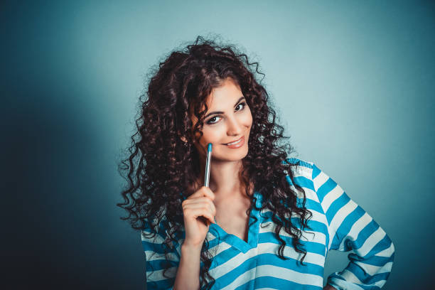 think. portrait happy beautiful woman thinking looking at you camera smiling pen on cheek isolated blue wall background with copy space. human face expressions, feelings body language imagination - stripped shirt imagens e fotografias de stock