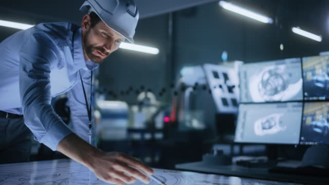 Industry 4.0 Modern Factory Office Meeting Room: Handsome Male Engineer Wearing Hardhat, Uses Pen on Touchscreen Digital Table to Correct, Draw Machinery Blueprints. High-Tech Electronics Facility